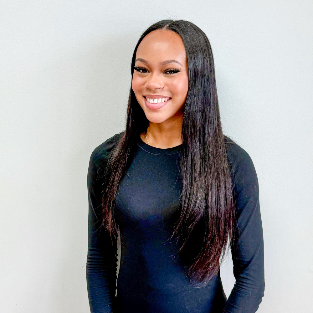 Portrait of a smiling woman with long straight black hair, wearing a black top, standing against a light gray wall.