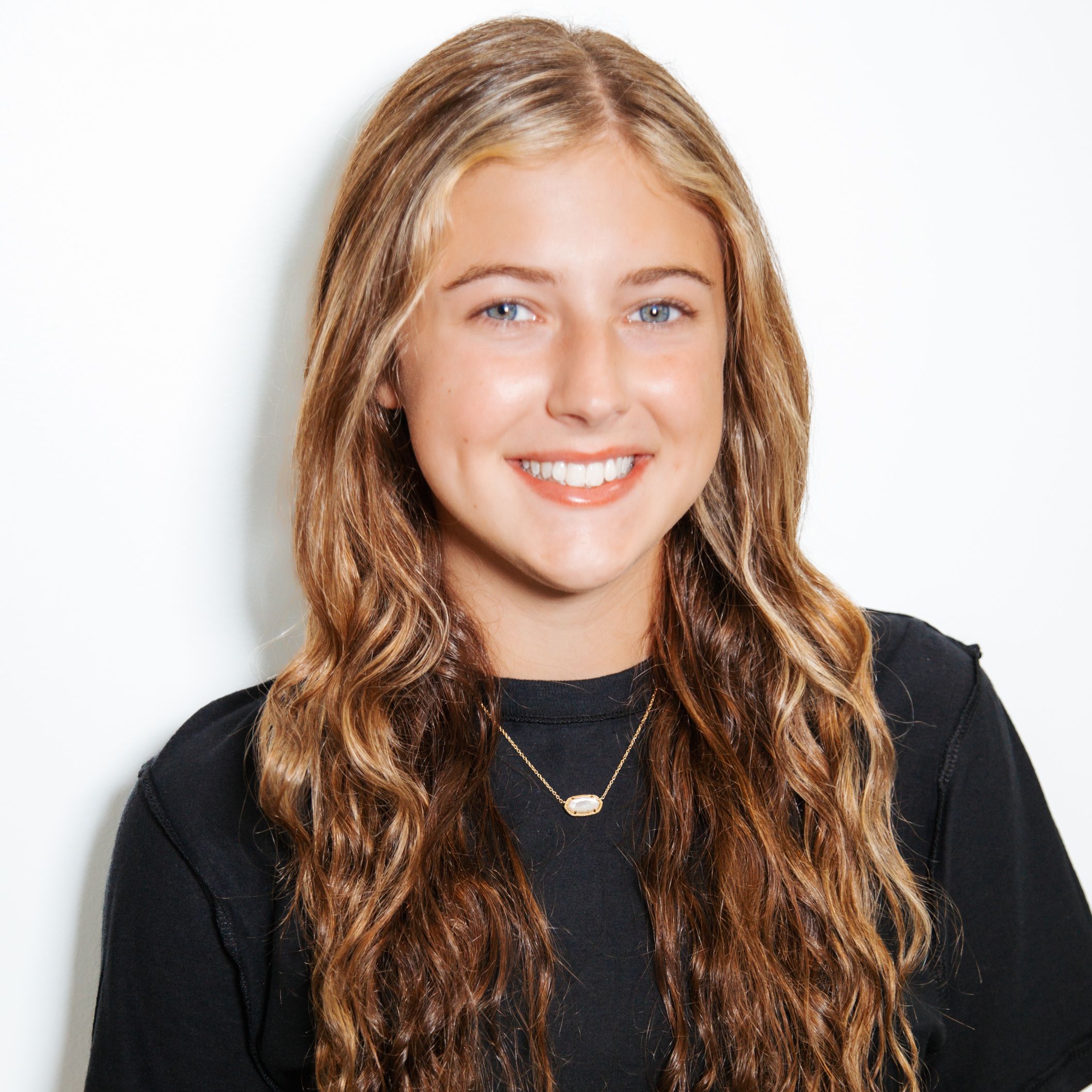 Smiling woman with long wavy brown hair wearing a black top and a delicate necklace, against a light background.