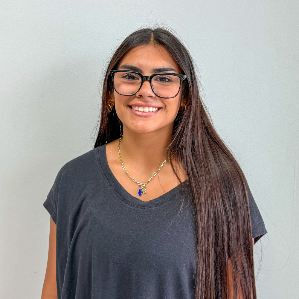 Smiling woman with long dark hair, black glasses, a black t-shirt, and a gold chain with a blue pendant.